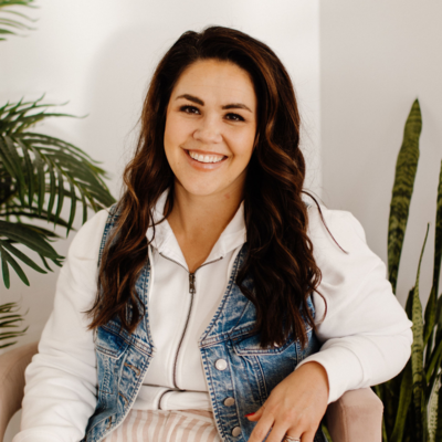 Woman with dark hair with white sweatshirt and jean vest sitting on pink chair with plants behind her