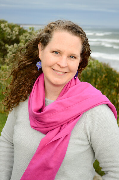 Sheila standing in front of the ocean wearing a pink scarf smiling