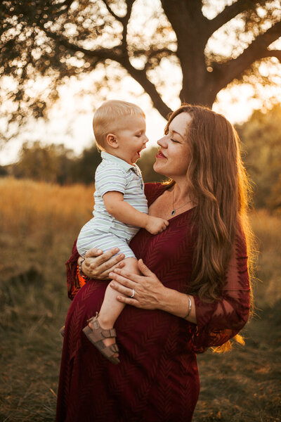 Mother with Toddler on arm. Standing in a meadow at sunset. Looking at each other. Little boy making faces at his mother. Mother genuinely smiling at him. Mommy and me photo.  Family Photography.