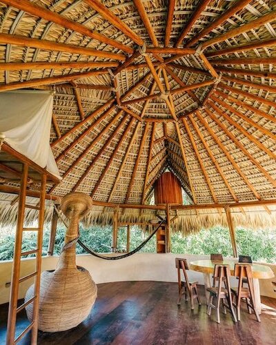 A spacious, open-air palapa at Barbarenas retreat in Puerto Escondido featuring a woven hanging chair, hammock, natural wood floors, and a round dining table—all surrounded by lush tropical greenery.