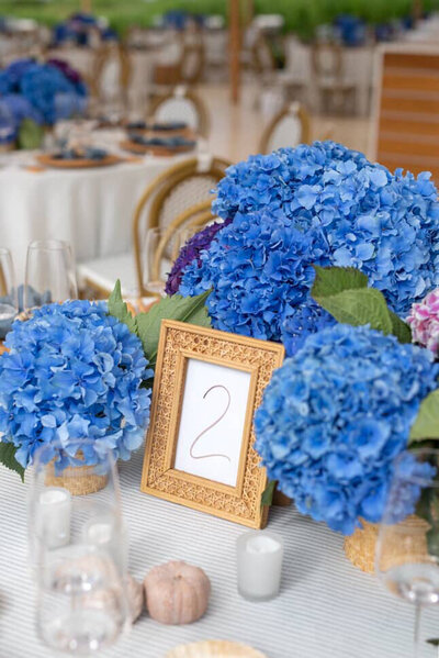 Hand lettered white table number in rattan picture frame with blue hydrangea flower arrangements for coastal wedding in Quogue, New York.