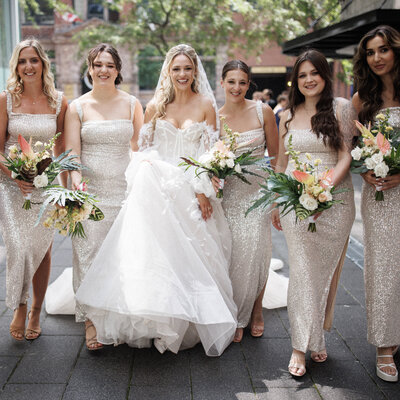 A group of bridesmaids holding exotic bouquets circle a bride in front of the Cathcart. 