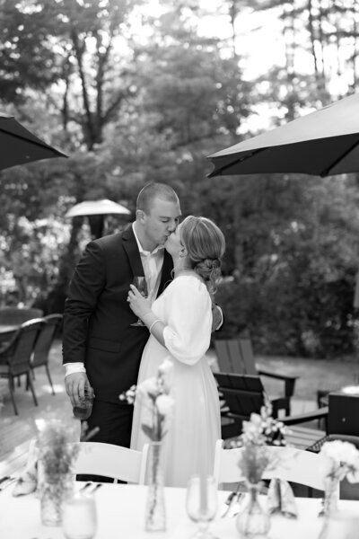 A couple sharing a kiss at an outdoor wedding reception. The bride, wearing a white dress, has her hair in an updo, and the groom is in a black suit. They are surrounded by tables with floral arrangements and large umbrellas, with trees in the background.