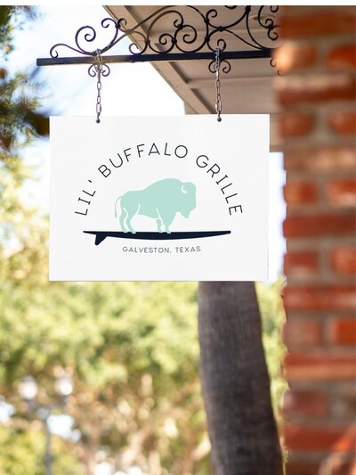 Outdoor hanging sign for Lil' Buffalo Grille with mint green buffalo and surfboard logo on a white background, mounted on a decorative iron bracket.