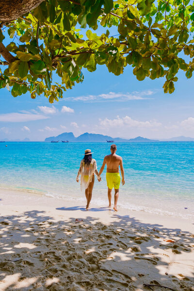 Couple holding hands while walking toward the ocean under leafy shade on a sunny beach.