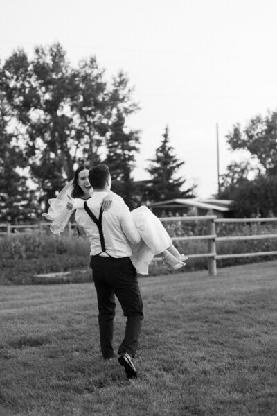 groom carrying bride across the grass while she gives a thumbs up