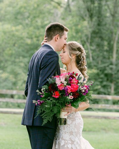A groom kisses his bride's forehead in a filed at Mayowood Stone Barn in Rochester following their Minnesota wedding. She is holding a bouquet full of pink and fuchsia flowers.