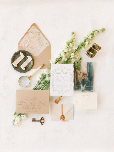 A decorative envelope with floral lining sits open on a white surface. Beside it are white flowers, a magnifying glass, and a metal tray with scrolls.