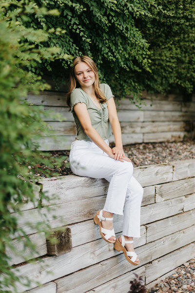 Senior girl in a green top and white pants sitting on a wooden wall in front of greenery
