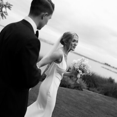 Black-and-white photo of bride and groom walking in the grass with the bay in the background at their East Coast wedding.