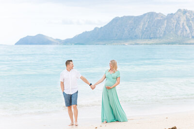 Expectant parents walking on the beach in a teal dress.