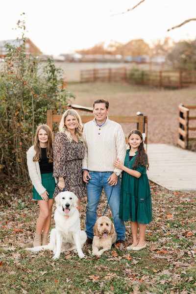 Family of four smiling with two dogs surrounded by fallen leaves.