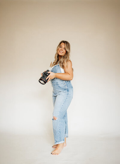 Full-body portrait of Charleston photographer Taylor Ragusett smiling and holding a camera in a light studio setting.