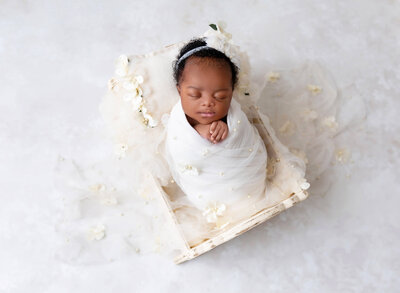 Baby girl is swaddles in a white wrap embellished with tulle and pearl detail, Her hands are peeking out of the wrap and folded under her chin.