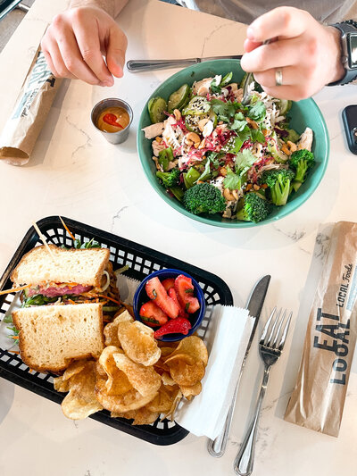 Image from above of a bowl of salad and a basket with a sandwich and chips on a table