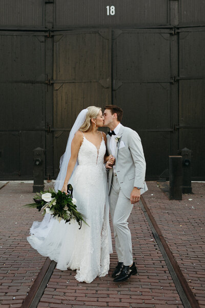 Bride and groom share a kiss in front of a large black door at Steam Whistle Brewing in Toronto. The bride, with a bouquet in hand and veil cascading over her white gown, stands beside her groom in a gray suit—captured by Toronto wedding photographer, Jennifer van Son.
