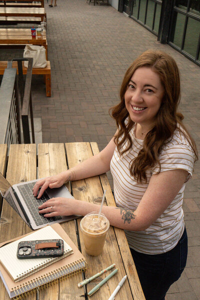Virtually Robyn, a woman and virtual assistant, with long brown hair smiles at the camera while working on a laptop at an outdoor wooden table. On the table are a phone, notebook, pens, and an iced coffee.