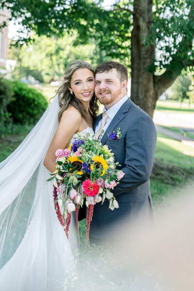 Bride and groom pose with a colorful bouquet under a tree. A summer wedding shot by Jackson Ms Wedding Photographer.