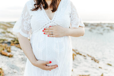 a pregnant woman in a white dress holds her baby bump while she stands on the sand dunes on Coronado beach near hotel del coronado