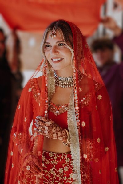A blonde haired bride wearing a red sari smiles brightly under a red canopy in Fairfax County Virginia
