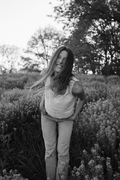 Cinematic wedding photographer leans towards camera with hands in her pockets and an infectious smile on her face, while standing in a field of golden flowers