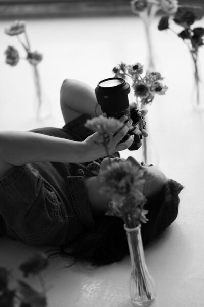 Black and white photo of a person lying on the floor, holding a camera, surrounded by flowers in vases. The scene is artistic and serene.