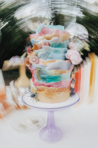 A stunning artistic wedding cake featuring rough-edged layers in watercolor shades of pink, teal, and orange, adorned with dried florals and set on a translucent lavender stand. The background features spinning reflections from disco balls. Captured by Dani Lacey Photographs.