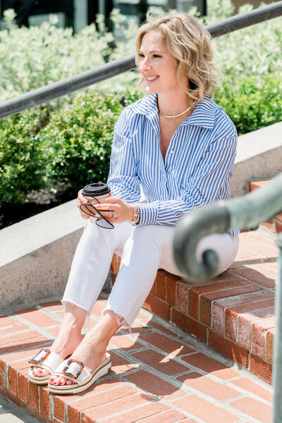 Julie Ladimer sitting outside her office on brick stairs wearing a blue and white striped shirt and white jeans
