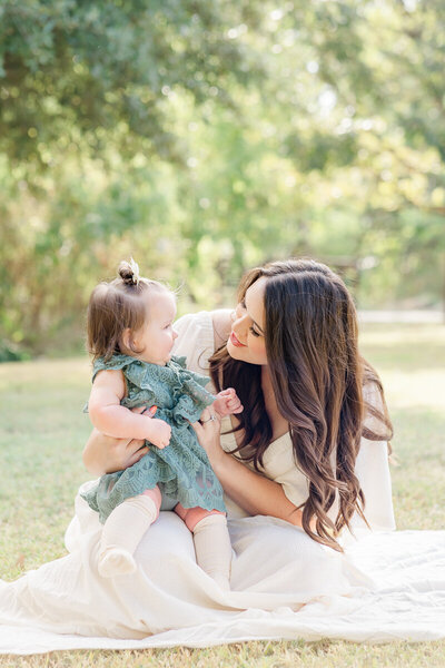 A mother sits on a white blanket with her baby girl on her lap at Katherine Fleischer Park. She looks at her baby smiling, in this tender shot taken by an Austin Family Photographer