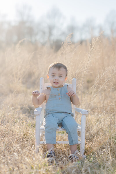 A toddler in blue overalls sits in a wooden chair in a field of tall grass at sunset