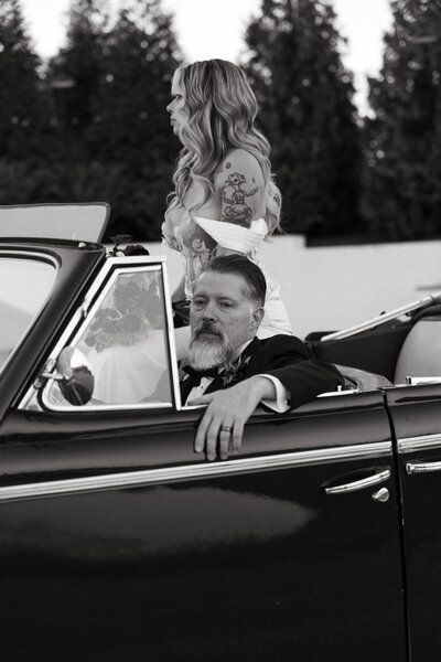 black and white wedding photograph by Photographer Knoxville TN with couple sitting in a vintage car, the groom at the steering wheel and his bride sitting on the back of the front seat so she can see over the windshield, they look different directions for a romantic wedding photo