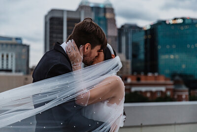 Shannon Lee, a Florida elopement photographer capturing a romantic kiss between a couple with the city skyline in the background and the bride's veil gently blowing in the wind.