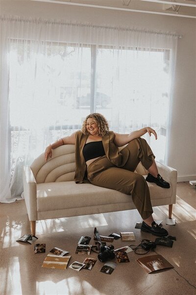 Andrea Cakmar, a wedding photographer, lounges on a modern, cream-colored sofa in a bright room. She is dressed in a tan pantsuit with a black crop top, surrounded by scattered photography prints and a camera on the floor.