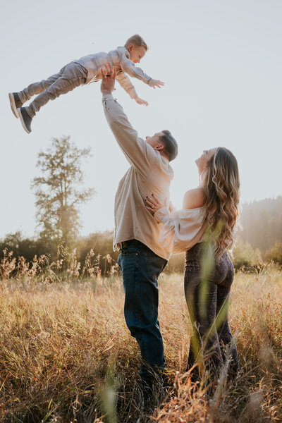 Father tosses son up in the air in a field with mom standing behind