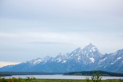 Panoramic view of the Grand Teton mountains with snow-capped peaks and a green valley below, showing a clear sky and serene landscape.