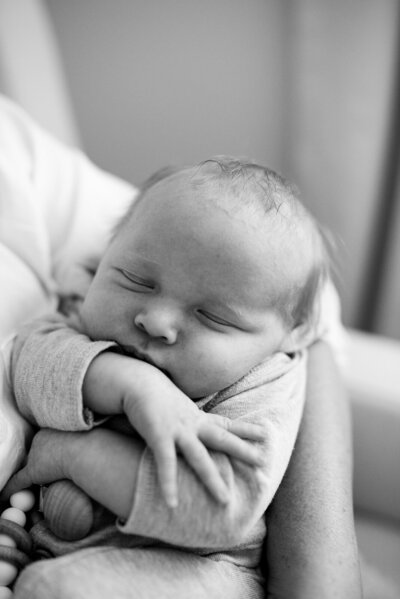 Baby held in parent's arms, snuggled in with his hands and arms wrapped over his chest sleeping