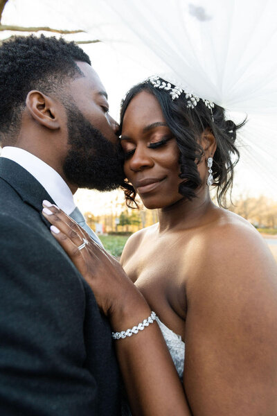 Couple in a warm embrace and the Groom is kissing his bride on the side of her face with the wind blowing her veil up over their heads.=