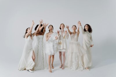 Six women in unique white bridal outfits reaching for bouquets mid-air in a playful wedding photoshoot
