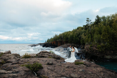 bride and groom standing on top of black rocks with a lake and trees in the background