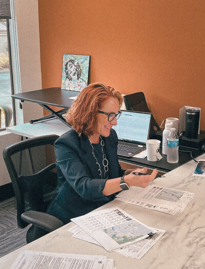 Carolynn Santaniello smiling at her desk, reviewing documents with a client in a modern office with an orange accent wall.