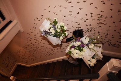 Marie carrying bunched bridal bouquets down the stairs