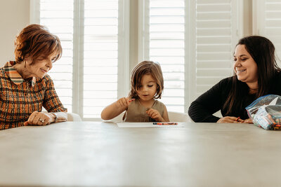 A young child colors with crayons at the kitchen table while their mother and grandmother look on, smiling and engaged, during a relaxed in-home lifestyle session in Scottsdale, Arizona.