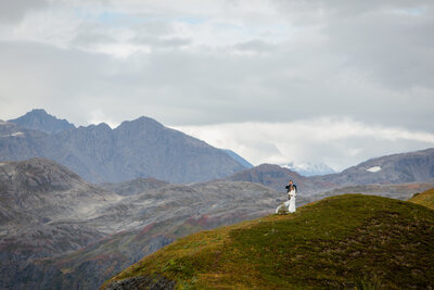 Newlyweds embrace on a mountain top  while miniaturized by the giant mountains around them.