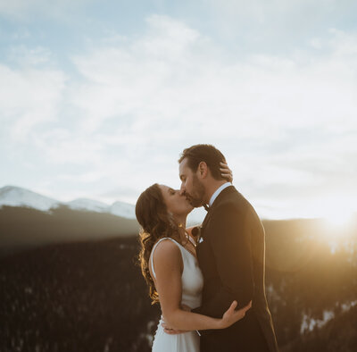 bride and groom are kissing as the sun sets. there are mountains behind them in colorado with snow on them. the trees are also in the background of the overlook.
