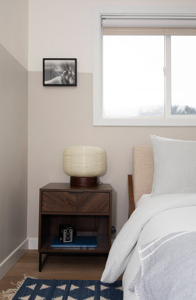 A corner of a bedroom showing part of a bed, a night stand with a lamp and a framed picture on the wall. Representing custom home interior design by Winsome Home Interiors.