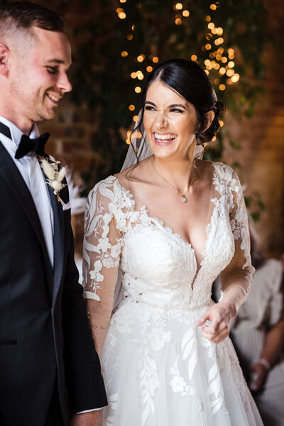 a bride and groom stood during their wedding ceremony at Shustoke Barn, bride is laughing and looking at her husband - photographed naturally and in the moment. Bride is wearing white lace and groom is in a black tie tux and smiling looking off camera