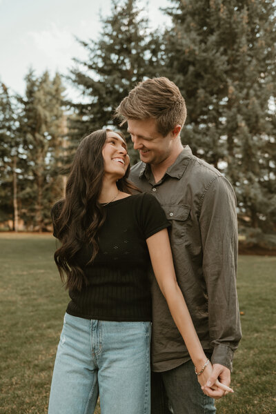 An engagement photo of a man and woman taken by Bailee Freeman Photography with green pine trees and a green grass field. Girl is in front of boy and looking back and up towards him while grabbing his hand. 