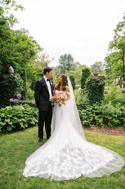 Bride and groom standing in a lush garden, holding hands and gazing at each other, with the bride's lace wedding gown flowing behind her.
