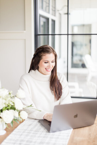 A woman with dark hair and a white turtleneck happily working on a laptop