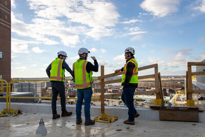 Three construction workers in safety gear discussing a project on a rooftop, overlooking a cityscape. Captured by Joanne Lee Photography, specializing in commercial and industrial photography in Maine